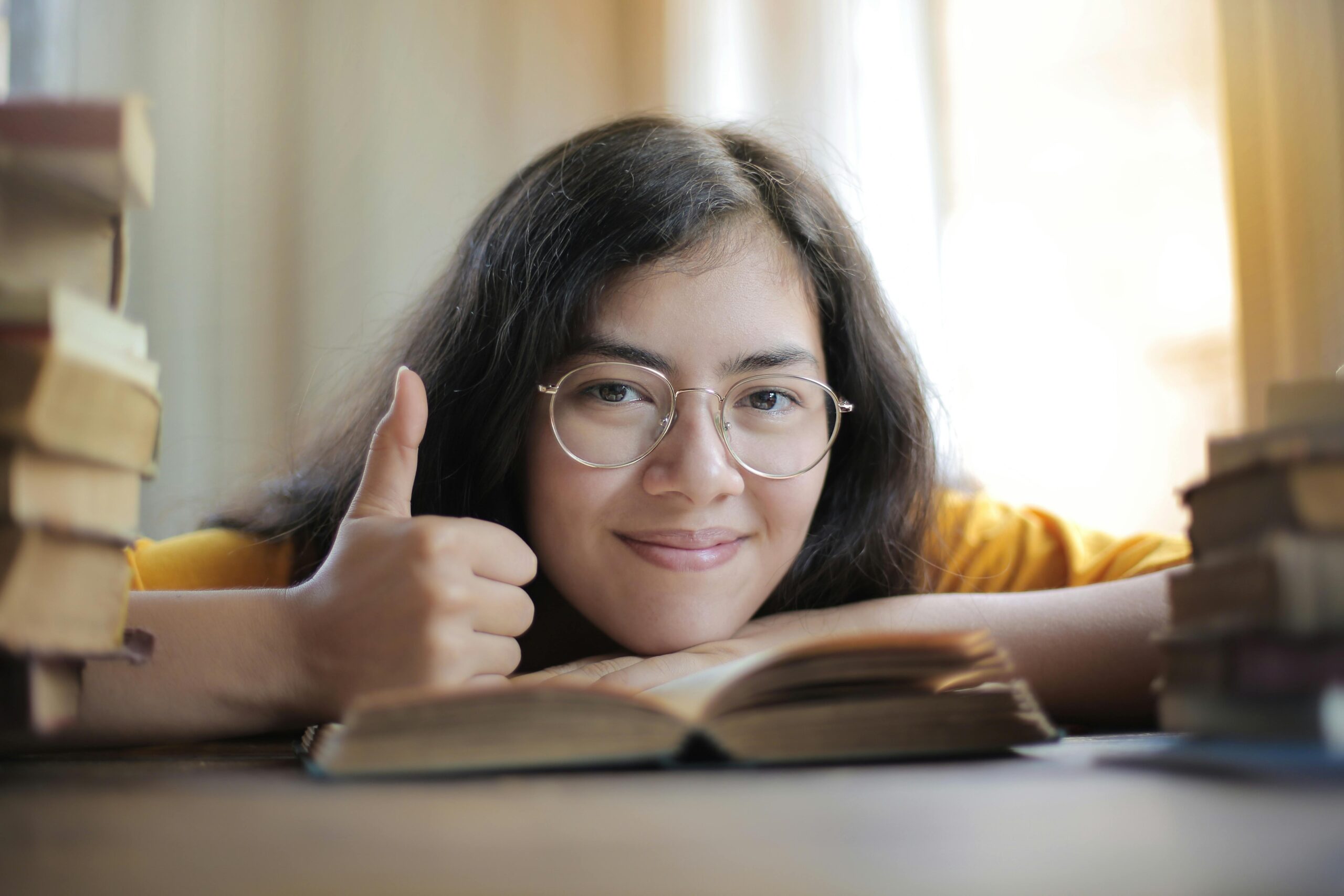 Portrait of a happy woman with glasses giving a thumbs up surrounded by books indoors.
